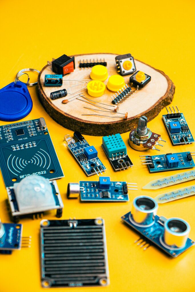 Colorful assortment of Arduino/Raspberry Pi compatible electronic components — sensors (PIR, ultrasonic, DHT, RFID-RC522), buttons, potentiometer, buzzer, capacitors, LEDs, and breakout boards — artistically arranged on a yellow background with a wooden slice in the center.