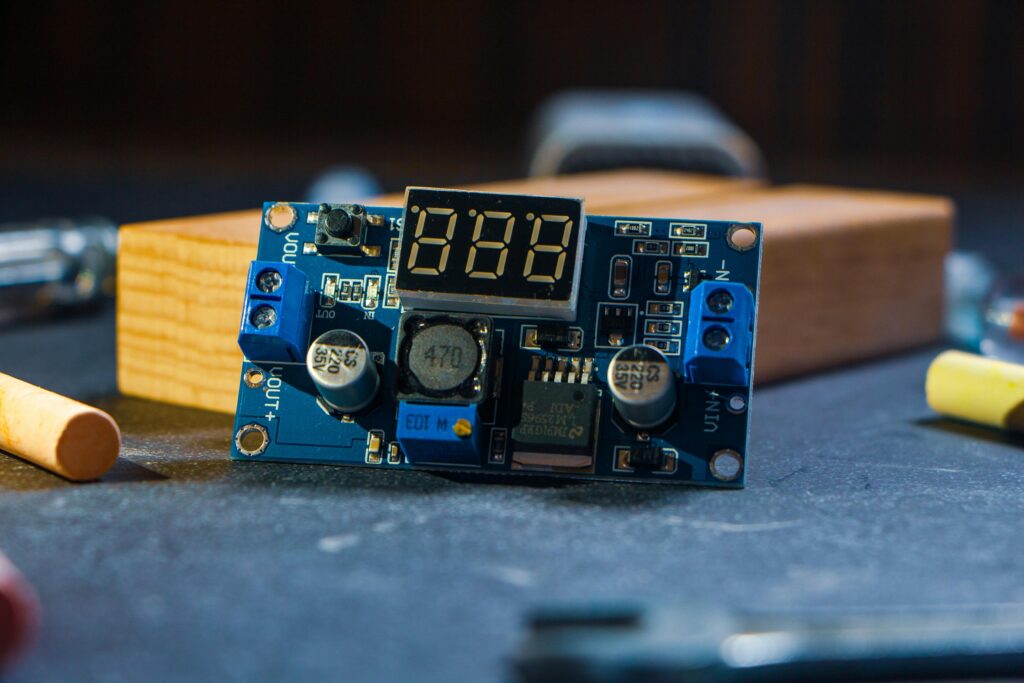 A small blue DIY electronics module featuring a 3-digit red 7-segment LED display showing '888'. The compact circuit board includes screw terminals labeled IN+ / IN− and OUT+ / OUT−, several electrolytic capacitors (one marked 220µF 35V, another 470), an adjustable voltage regulator IC (likely LM2596-based with 'ADJ' potentiometer), a small inductor, push buttons, and various SMD components. The module is placed on a wooden surface next to scattered cigarette butts and a wooden block in the background.