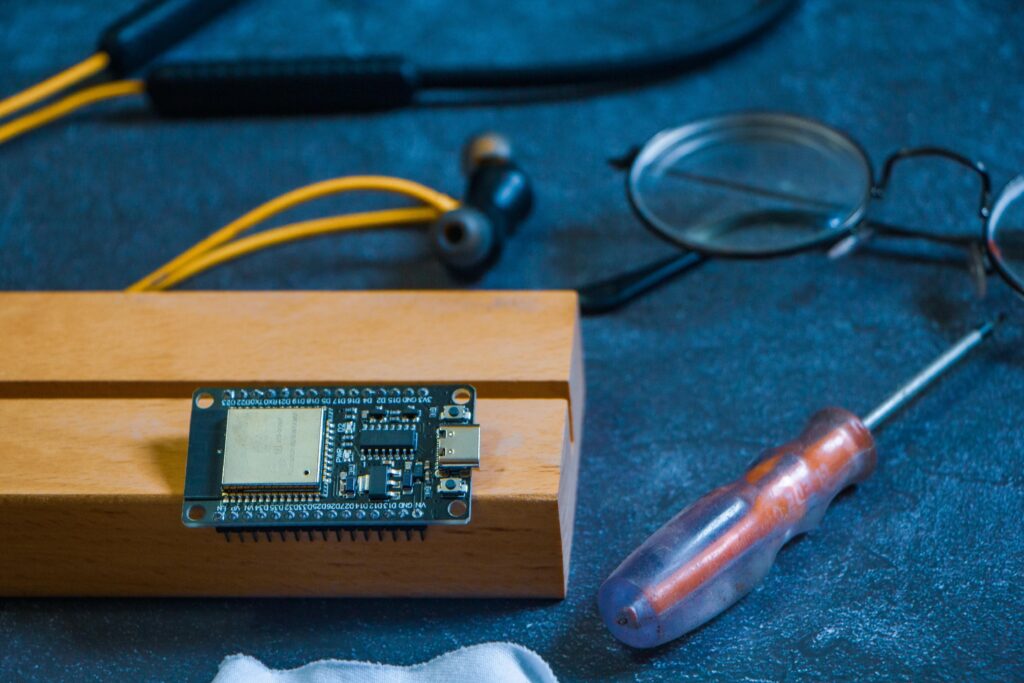"A compact black ESP32 development board (likely an ESP32-C3 or similar ultra-small variant) with a large square metal-shielded chip on top, USB-C connector, and two rows of pin headers, resting on a small wooden block. Surrounding it are a small flathead screwdriver, yellow-and-black wired earbuds, round reading glasses, and a stethoscope in the softly lit blue-toned background, creating a maker/workbench aesthetic.